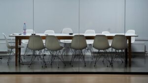 Modern conference room with a long wooden table and a combination of white and taupe Eames-style chairs reflected in a glass partition, a solitary water bottle and notepad on the table indicating a recent or upcoming meeting.